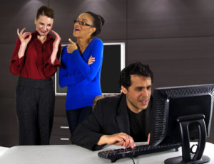 Mann jobber ved en PC, mens to kvinnelige kollegaer står bak ham og prater.