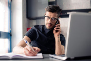 Mann sitter ved et skrivebord med en PC og noterer i en bok mens han snakker i telefonen.